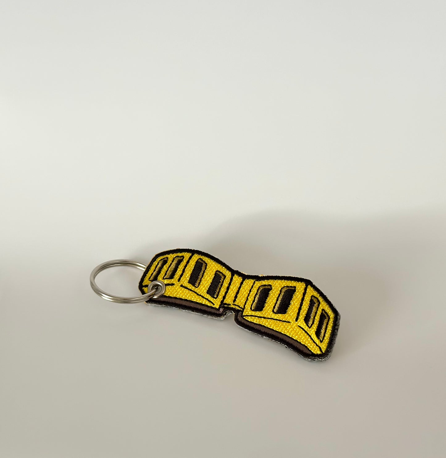Yellow and black goggles/glasses embroidered keychain on a light gray background that a certain hero wears 