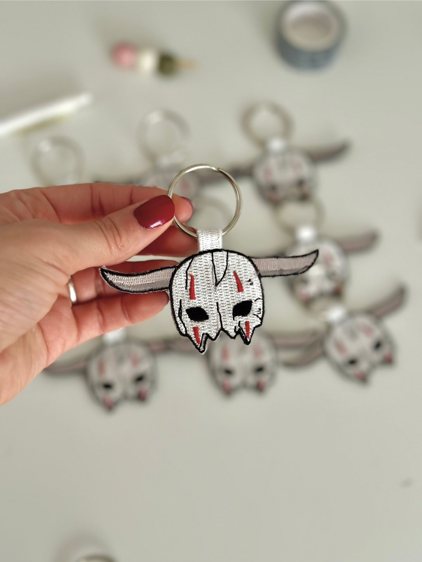 Hand holding a keychain with a skull mask that belongs to a blue dragon warrior against a blurred background of similar keychains.
