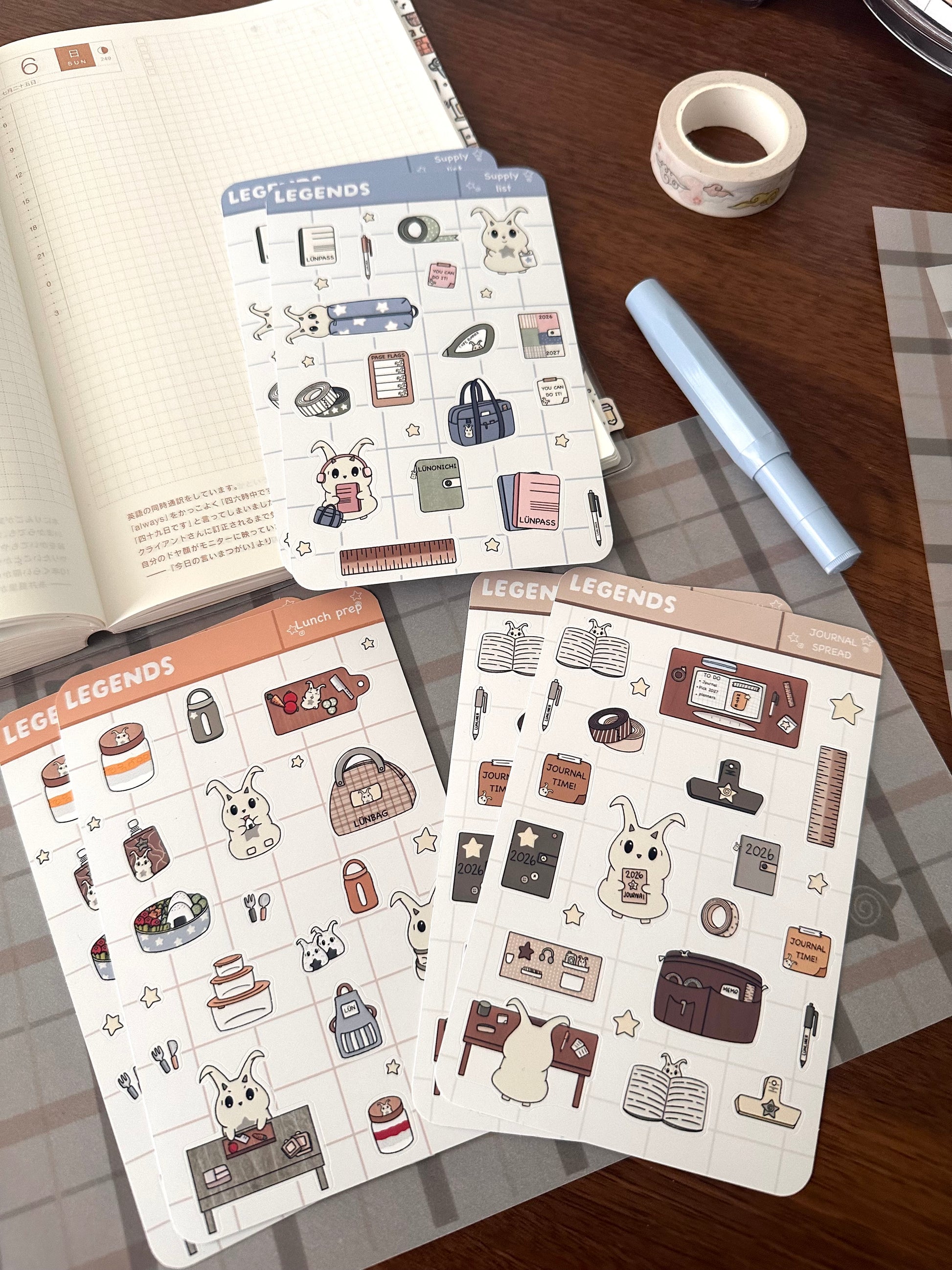 Three decorative planner sticker sheets with lün in our lunch prep, back to supplies, and journal spread stickers are arranged on a wooden desk alongside an open notebook, washi tape, and a light blue pen.