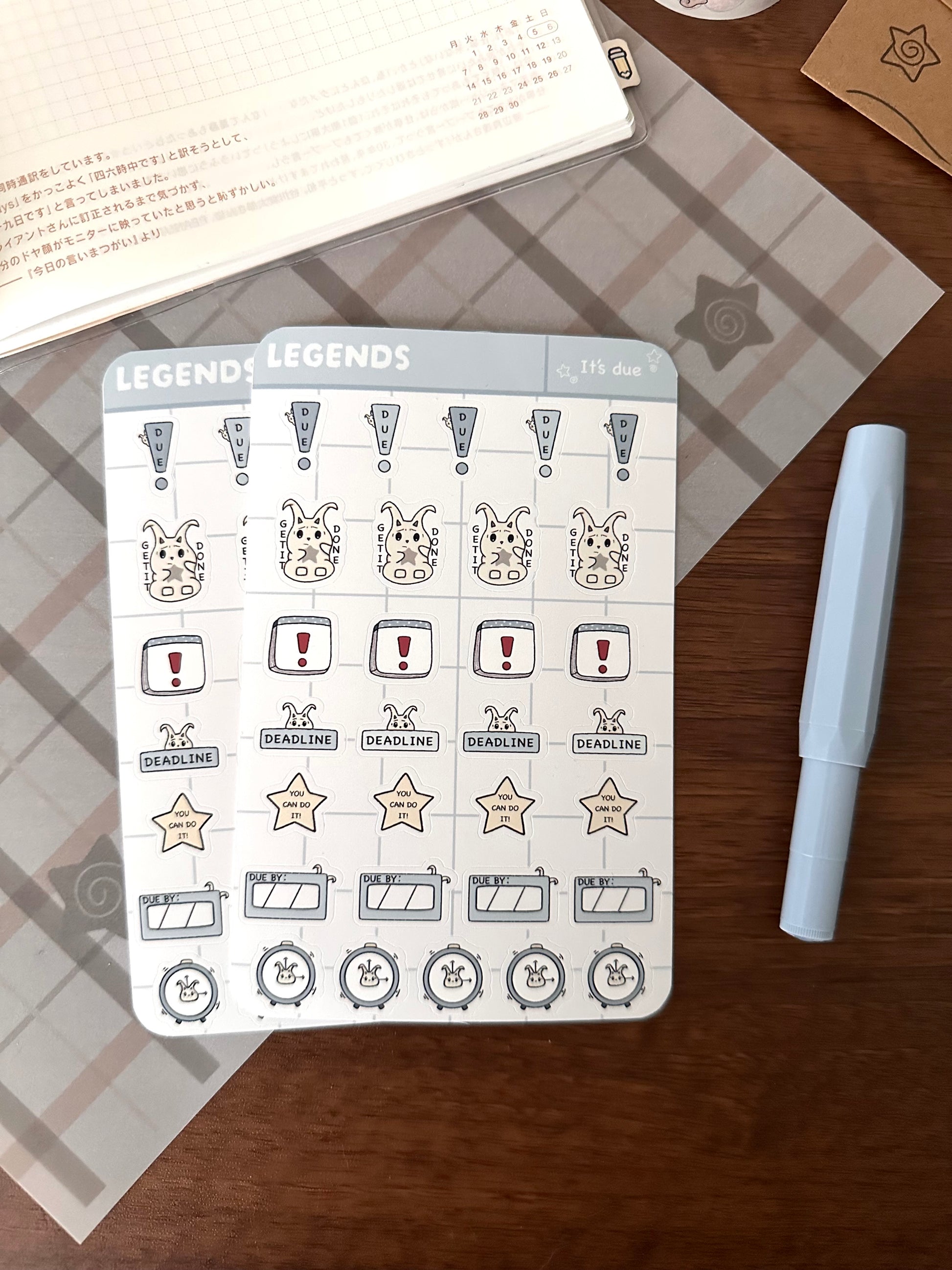 Two planner sticker sheets with original character lün and alarm clock illustrations, displayed on a wooden desk next to a light blue pen.