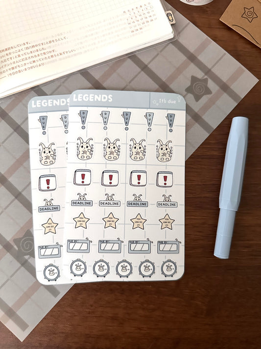 Two planner sticker sheets with original character lün and alarm clock illustrations, displayed on a wooden desk next to a light blue pen.