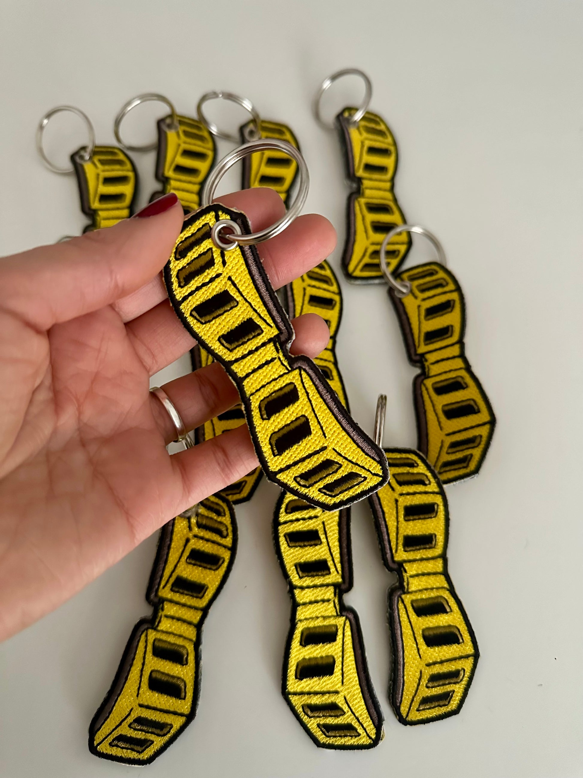Yellow and black goggles/glasses embriodered keychain that a certain hero wears, with my hand holding one.