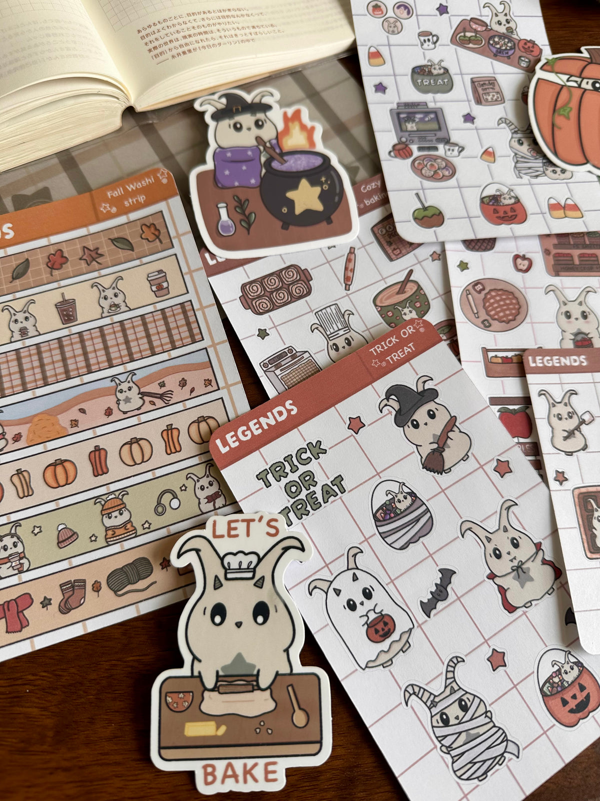 A collection of cute Halloween-themed planner stickers and washi tape sheets arranged on a wooden table next to an open book. The designs feature lün in various Halloween roles like a witch, baker, ghost, and mummy, along with pumpkins, cauldrons, and spooky legends.