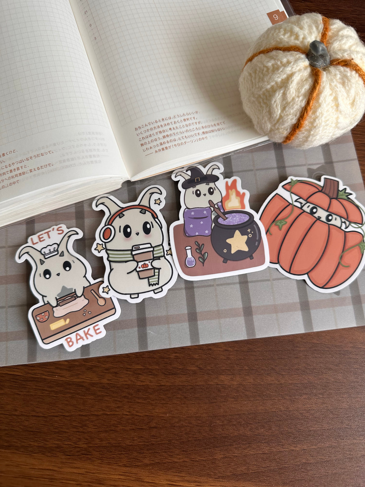Four cute Halloween-themed die-cut stickers displayed on a plaid mat, with an open notebook and a small yarn pumpkin in the background.