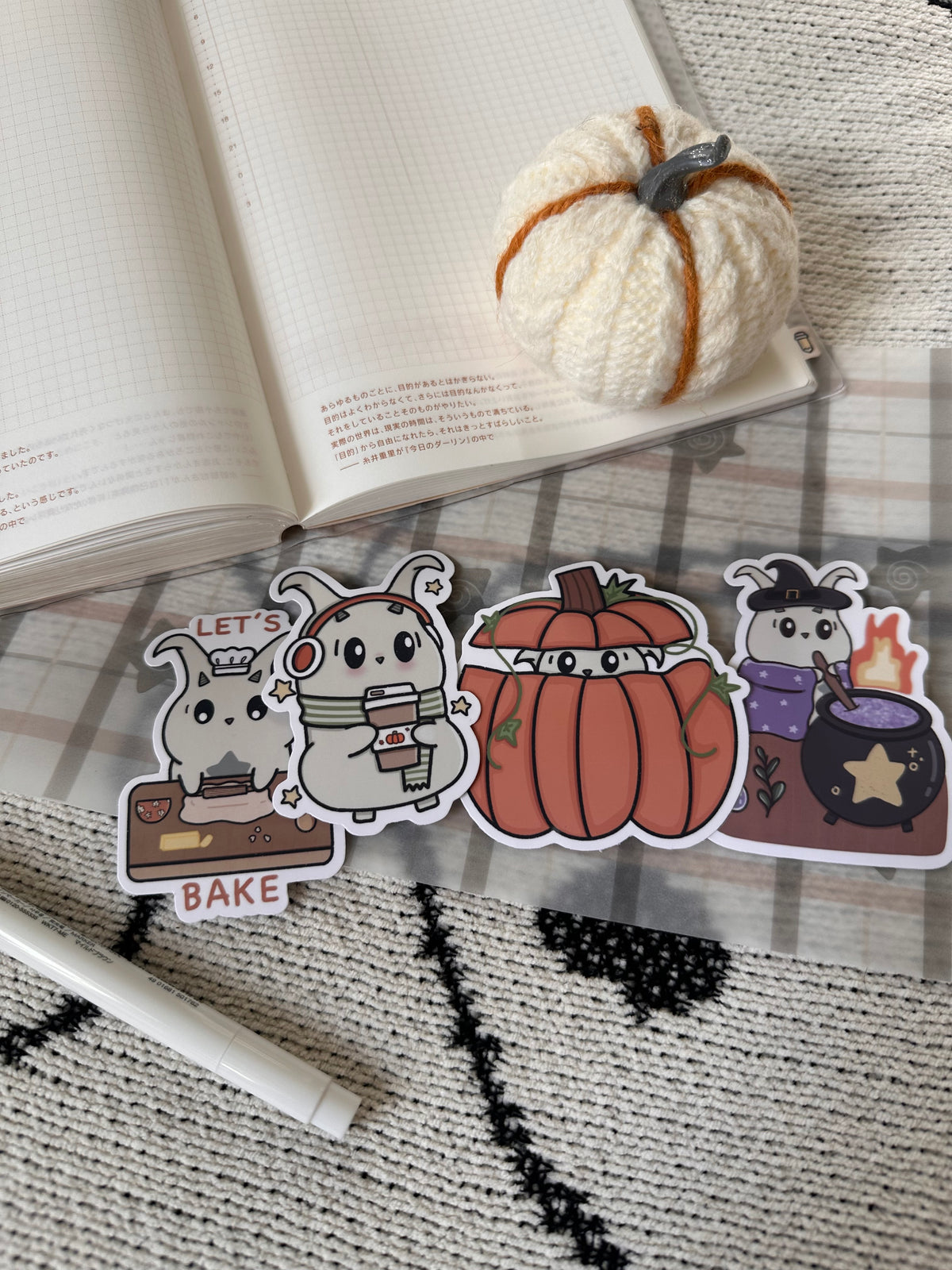 Four cute Halloween/ fall -themed stickers featuring lün baking, a pumpkin face, and a witch by a cauldron, arranged on a plaid blanket with an open notebook and a small knit pumpkin prop in the background.