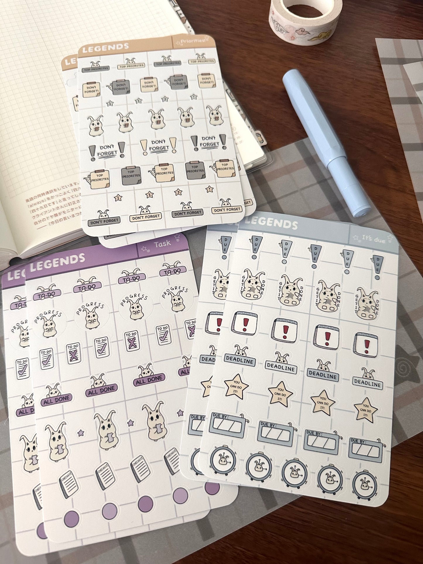 Three functional planner sticker sheets with lün in our todo list, deadlines and priorities stickers are arranged on a wooden desk alongside an open notebook, washi tape, and a light blue pen.