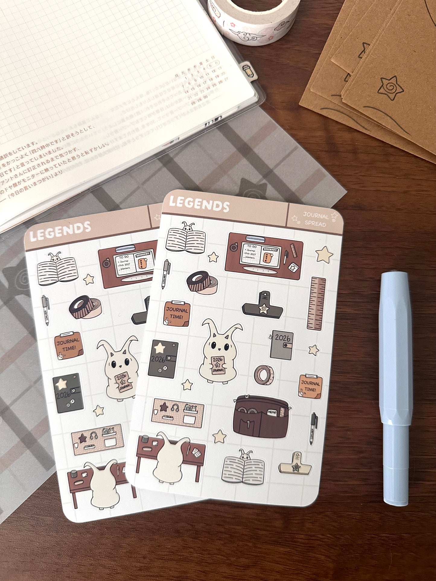 Two stickers sheet that have Lün’ doing journal stuff and journal supply stickers. On top of a wooden desk next to a blue pen 
