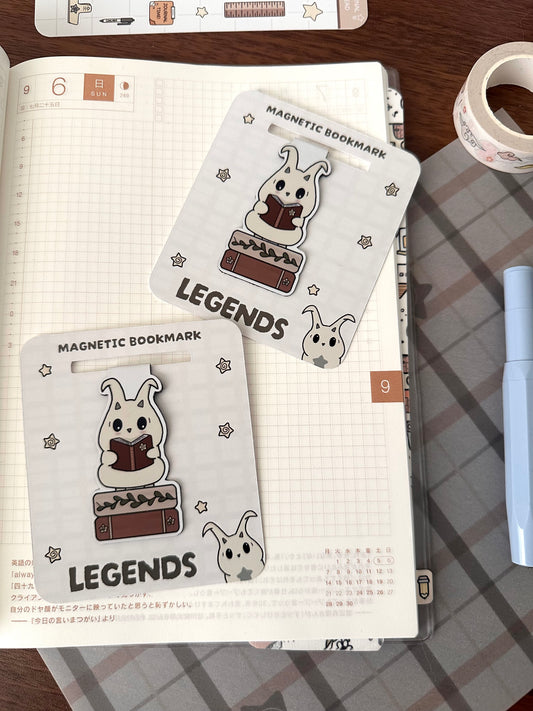 Two book themed magnetic bookmarks with original character lün reading and standing on two books. Bookmarks are laying flat on an open planner. 
