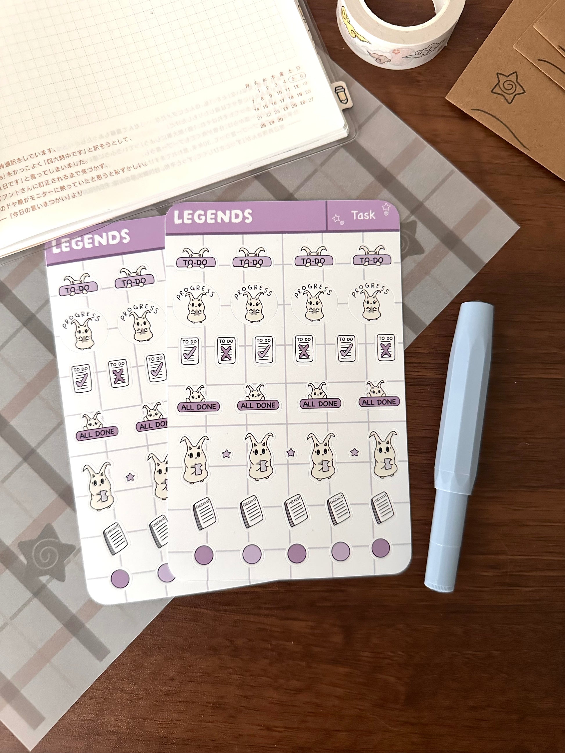 A planner sticker sheet labeled with lün and task icons, styled on a wooden desk with a light blue pen, washi tape, and a notebook.