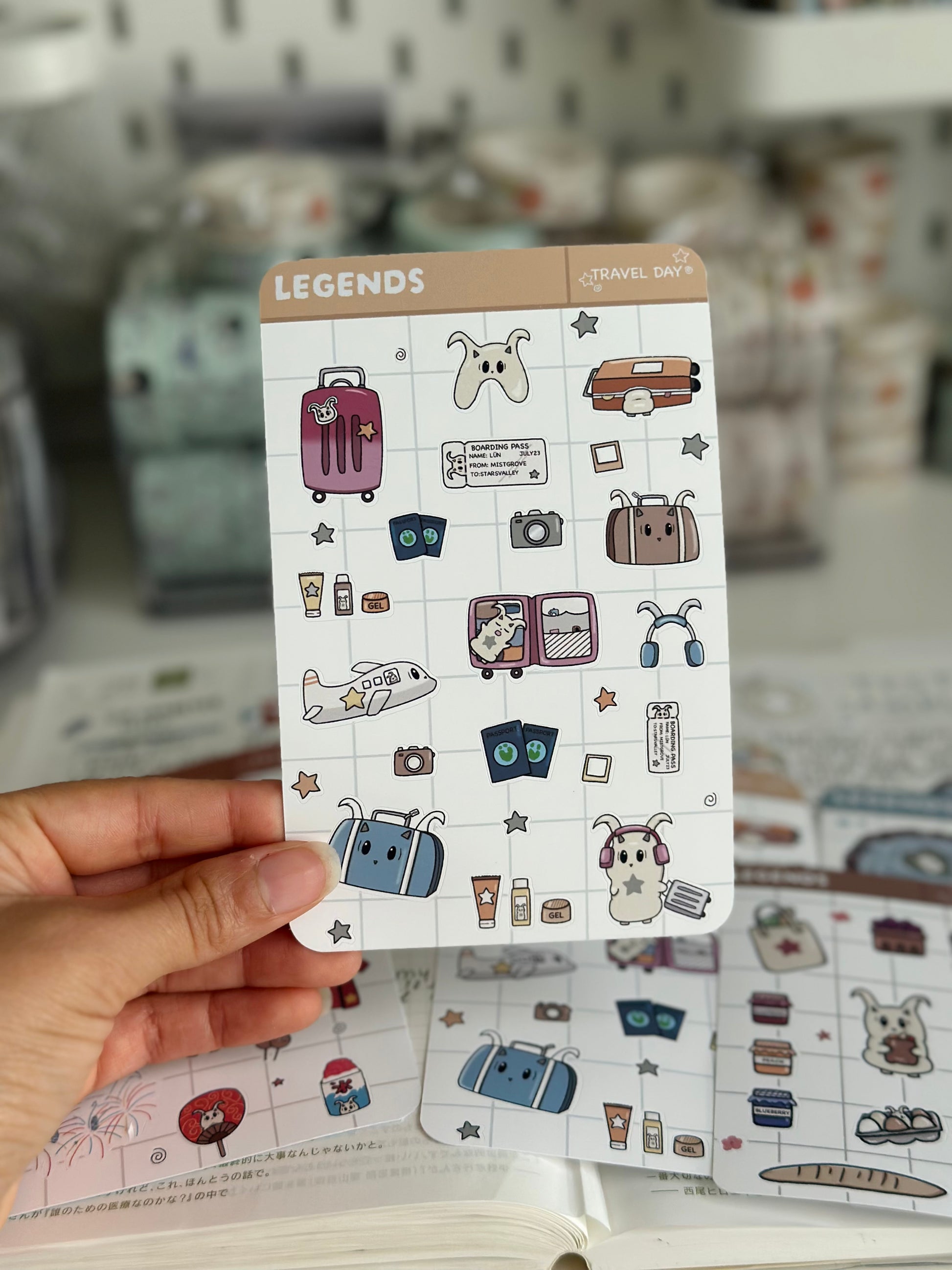 My hand holding the travel day sticker sheet that has a suitcase, minis, and passports on it, hovering over my journal. 